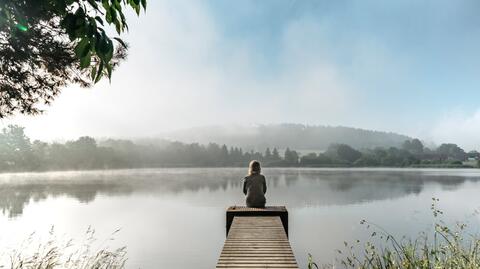 Stimmungsbild zum Internationalen Tag der Selbstfürsorge 2025, das eine Frau zeigt, die an einem See sitzt